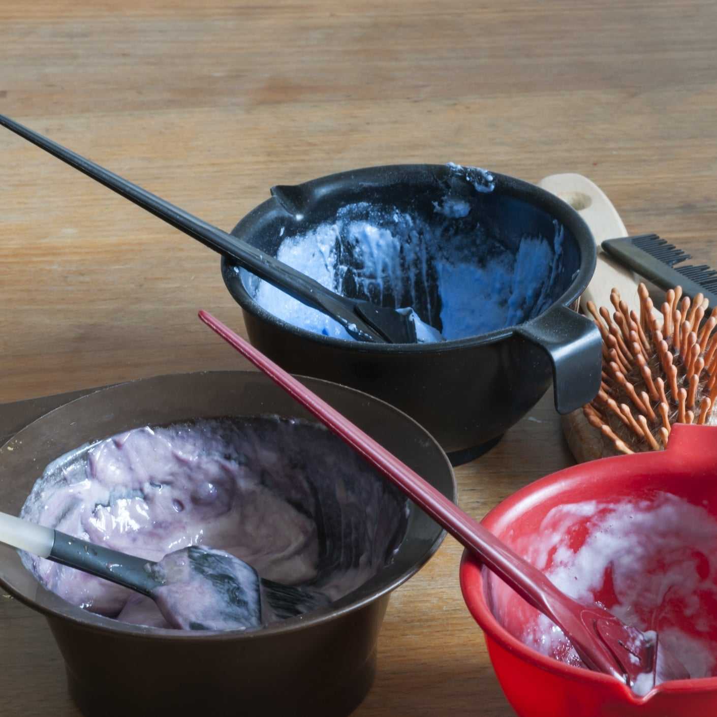 Hair dyeing tools and color bowls with brushes on a wooden table.